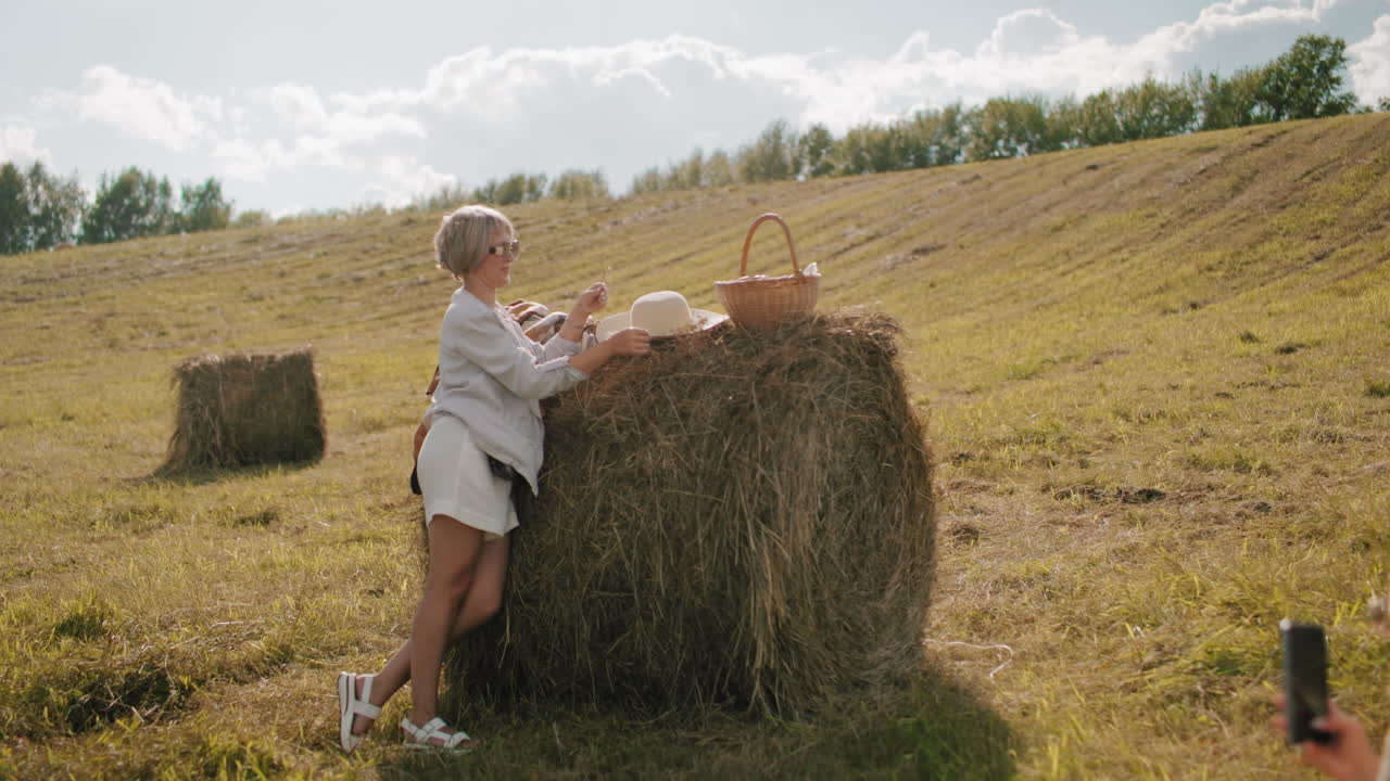 женщина, стоящая на коленях в открытом поле, фотографирует стильного друга, опирающегося на пучок сена, регулирующего шляпу возле пикника. золотой солнечный свет отбрасывает теплый свет на сельский пейзаж.