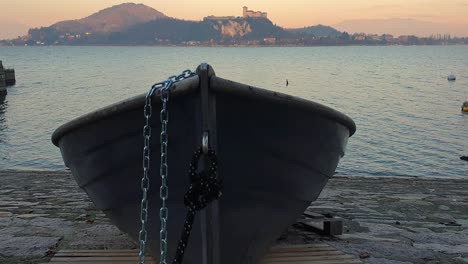 Barco-De-Pesca-Encallado-Y-Atado-Con-Una-Cadena-De-Hierro-Frente-Al-Lago-Maggiore-Y-El-Castillo-De-Angrya,-Arona,-Italia