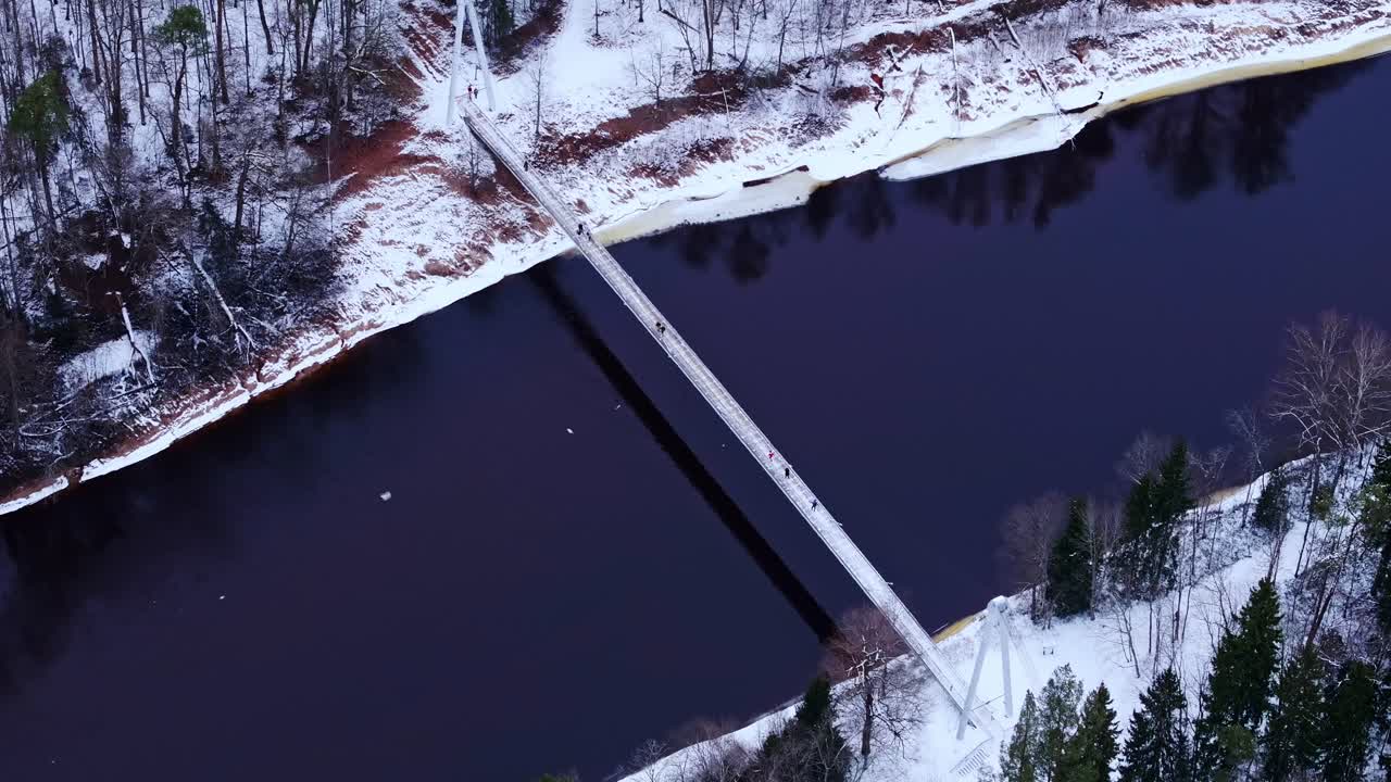 Suspension footbridge above dark river framed by snowy forest in Latvia