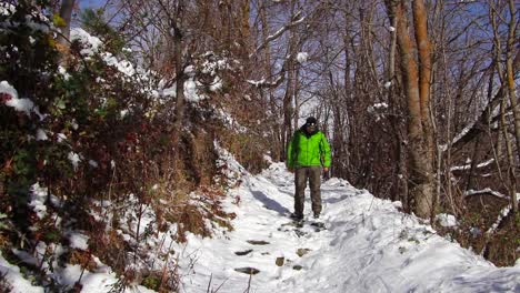 Mann-In-Einem-Grünen-Anorak,-Der-Einen-Bergpfad-Entlang-Geht,-Der-Zwischen-Bäumen-Und-Büschen-In-Einem-Verschneiten-Wald-Verläuft-Und-Hinuntergeht