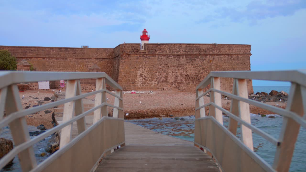 View from a floating walkway or bridge leading to Fort de Brescou with red lighthouse, under soft afternoon light in Cap d'Agde, France