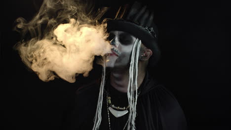 sinister man with professional skull makeup exhaling cigarette smoke from his mouth and nose