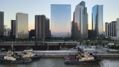 Carro-Aéreo-A-La-Izquierda-De-Los-Barcos-En-Los-Muelles-De-Puerto-Madero-Cerca-De-La-Autopista-Paseo-Del-Bajo-Y-Los-Rascacielos-Al-Atardecer,-Buenos-Aires