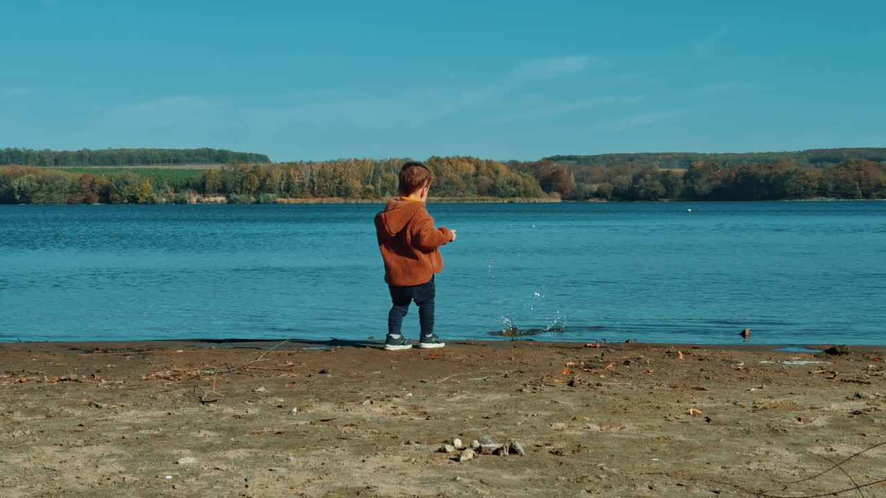 Cute Caucasian toddler playing on the river bank in autumn. Little boy picks a stone and takes it to the water.