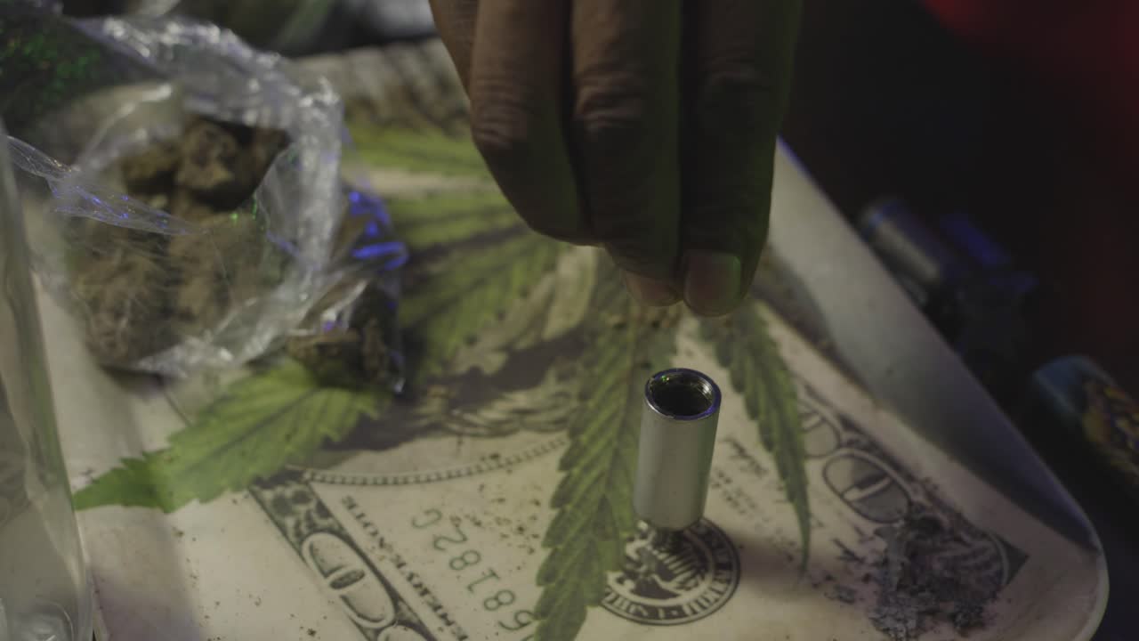 Close-up of a hand packing cannabis into a bowl on a tray featuring a dollar bill and marijuana leaf graphic.