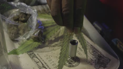 close-up of a hand packing cannabis into a bowl on a tray featuring a dollar bill and marijuana leaf graphic.