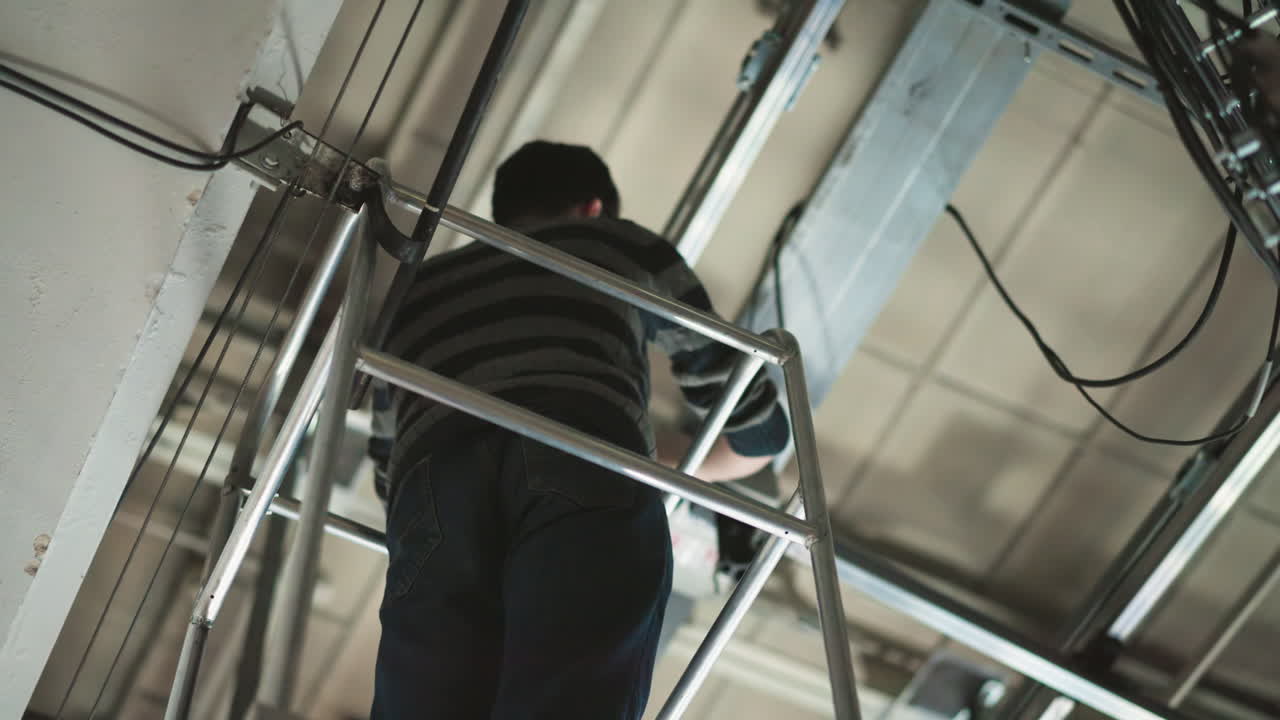 Person working at height, standing on ladder, adjusting equipment mounted on ceiling grid, industrial setting, cables visible, wearing sweater and jeans, maintenance or installation task