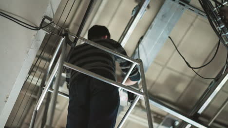person working at height, standing on ladder, adjusting equipment mounted on ceiling grid, industrial setting, cables visible, wearing sweater and jeans, maintenance or installation task