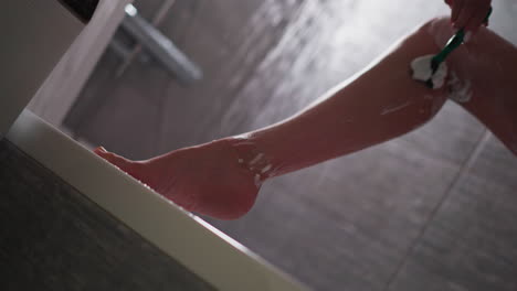woman removes hair from leg with razor in shower closeup. lady shaves body with cream and modern blade in bathroom. hygienic and beauty routine