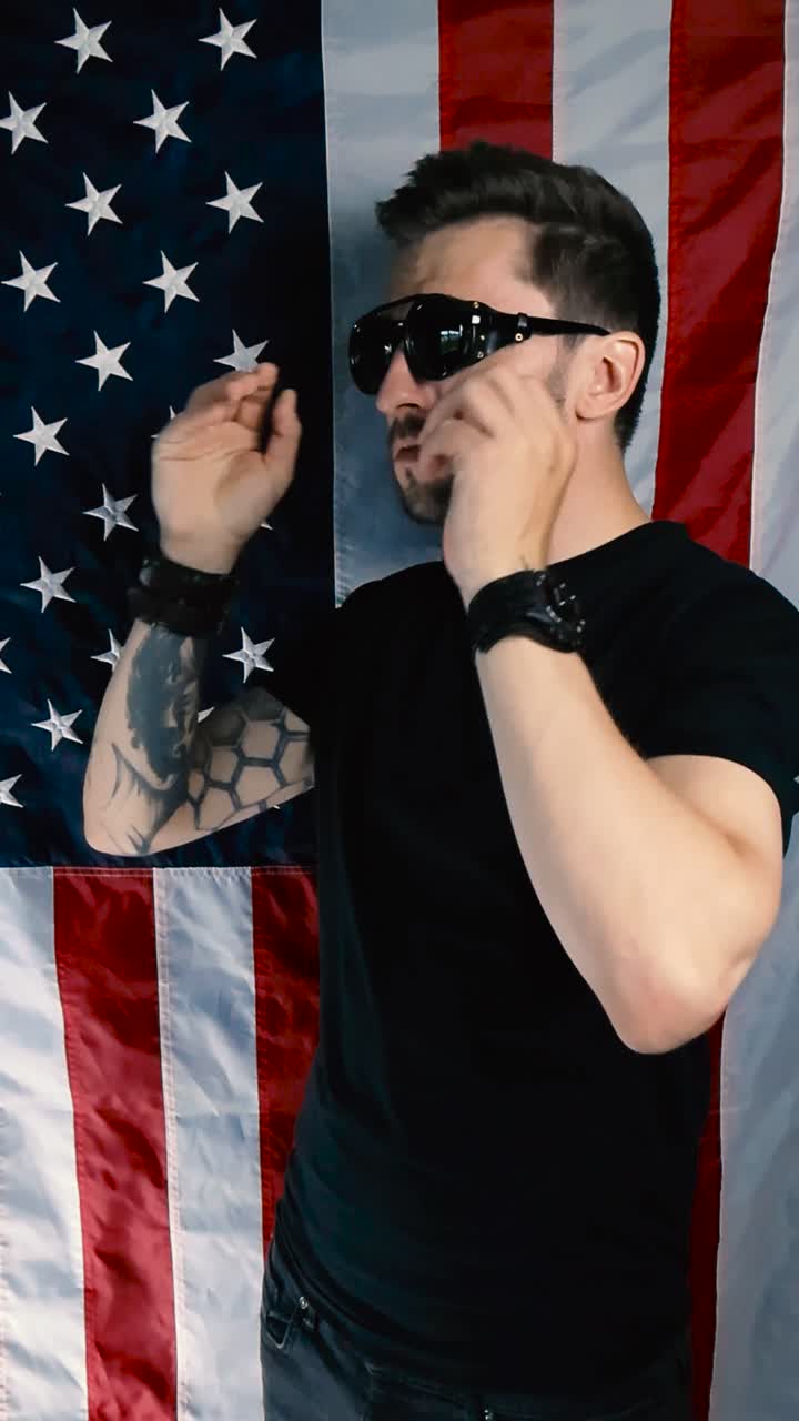 Tattooed man who is standing in front of a vertical American flag puts on retro and vintage goggles or sunglasses and shakes his head while arms are crossed, showing he does not like something