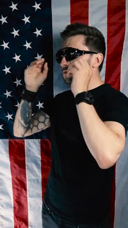 tattooed man who is standing in front of a vertical american flag puts on retro and vintage goggles or sunglasses and shakes his head while arms are crossed, showing he does not like something