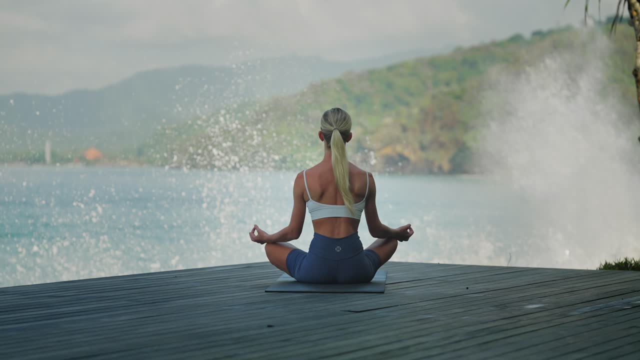 Athletic woman sitting on yoga mat doing meditation with huge crashing waves in background, Bali shore
