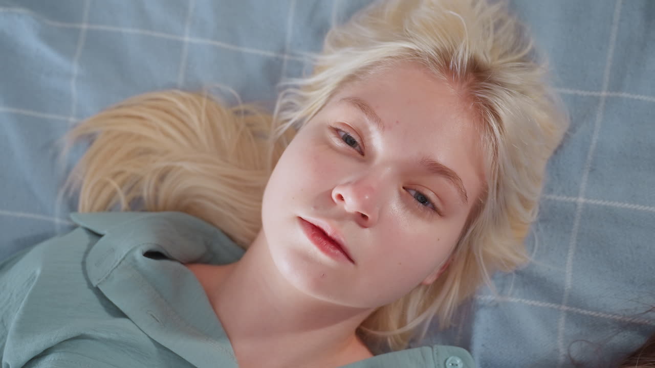 Close up of young blonde woman resting on bed with head turned slightly and eyes open, wearing teal shirt, lying on checkered blue blanket in soft natural lighting