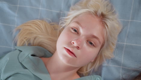 close up of young blonde woman resting on bed with head turned slightly and eyes open, wearing teal shirt, lying on checkered blue blanket in soft natural lighting