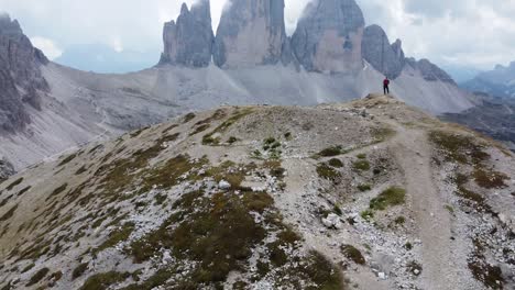 Una-Foto-Genial-De-Un-Dron-De-4k-De-Un-Excursionista-Parado-En-La-Cima-De-Una-Montaña-Rodeado-Por-Las-Impresionantes-Formaciones-Rocosas-Y-El-Paisaje-Montañoso-De-Los-Dolomitas-En-Italia---Mirando-El-Impresionante-Tre-Cime-Di-Lavaredo