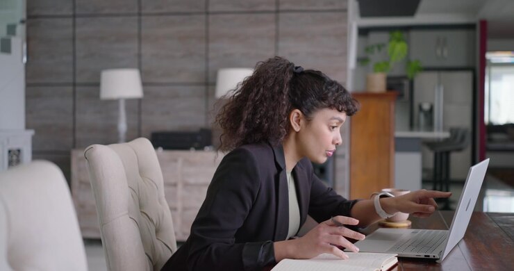 African American woman in blazer starting work dining table taking notes checking laptop