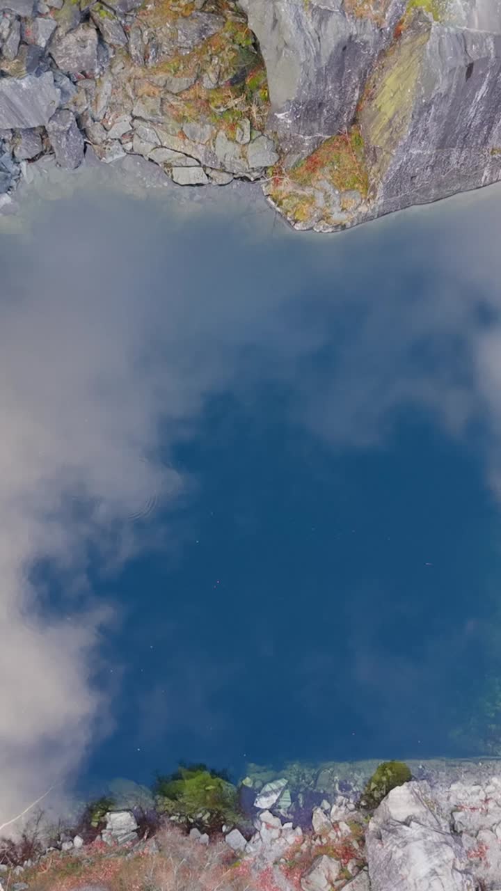 Ripresa aerea verticale ravvicinata dall'alto verso il basso che inizia con il cielo e il riflesso del drone nel lago della cava. Quando si alza, il suo riflesso si restringe e compaiono pareti di ardesia, rivelando l'illusione ottica di guardare verso il basso, non verso l'alto.