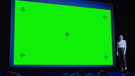 business conference stage: female keynote speaker presents new product to the audience, behind her movie theater with green screen, mock-up, chroma key. live event, tech device reveal