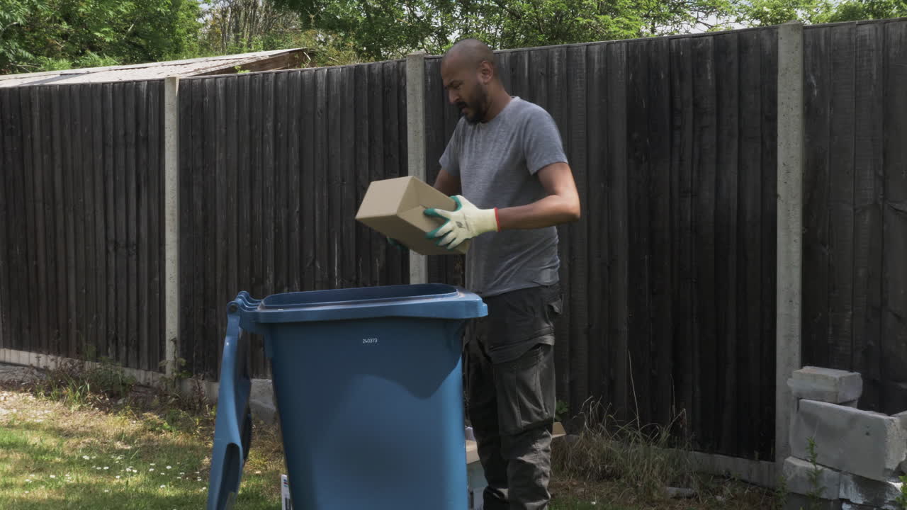 Adult UK Asian Male Sorting Cardboard Beside Blue Recycling Bin In Garden