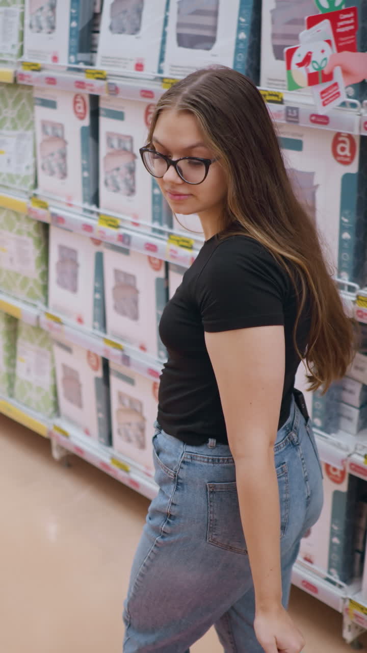 vista trasera de una mujer joven con camiseta negra y vaqueros caminando por el pasillo de la tienda, revisando estantes llenos de productos de cama mientras sostiene bolsas de compras, entorno minorista brillante