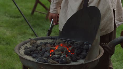 a revolutionary war reinacter blacksmith puts a piece of metal in hot coals to heat up while he pumps the bellows