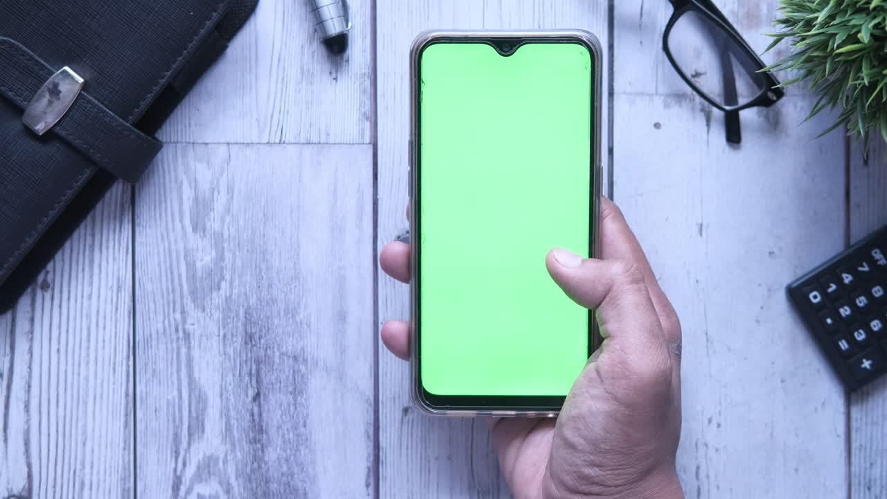 persona sosteniendo un teléfono inteligente con una pantalla verde en un escritorio de madera con accesorios