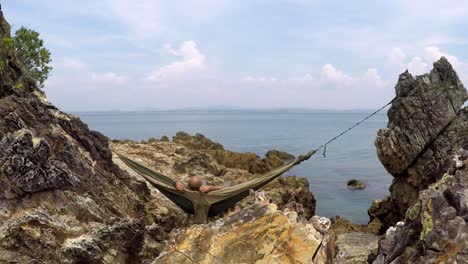 man relaxing in a hammock between rocks and looking at the sea