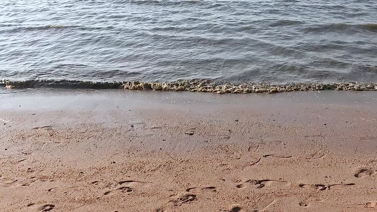 Footprints trail across the sandy beach as a lone boat floats on gentle waves under a clear sky.