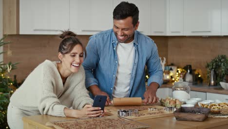 Hombre-Latino-Haciendo-Galletas-De-Jengibre-Y-Mujer-Caucásica-Usando-El-Teléfono-Junto-A-él.