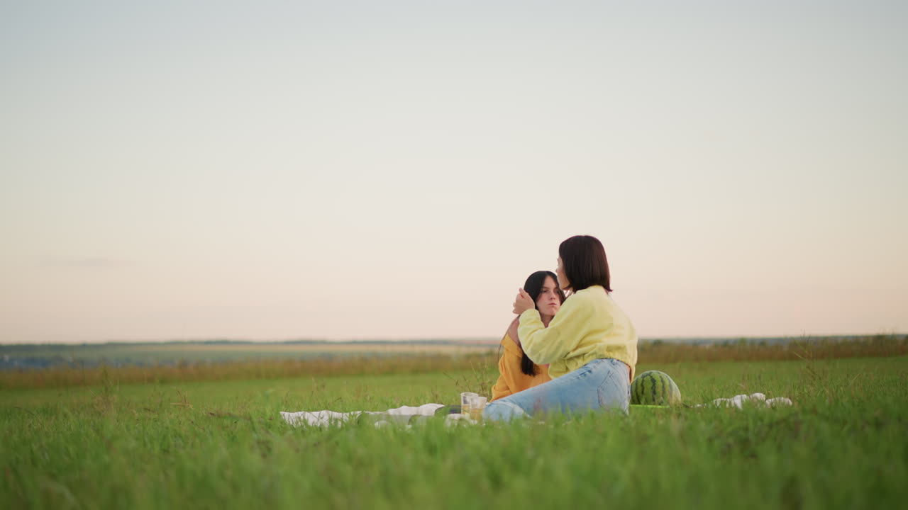 해 질 녘 목초지에서 피크닉을 즐기는 두 여성이 간식을 나눠 먹으며 웃고 있어요. 워크숍을 떠나 주말을 즐기는 직장 동료들이 담요 위에서 장난스럽게 서로 먹여주는 모습이 보입니다. 넓게 펼쳐진 목가적인 풍경과 부드러운 저녁놀, 캐주얼한 재킷과 데님 차림이에요.