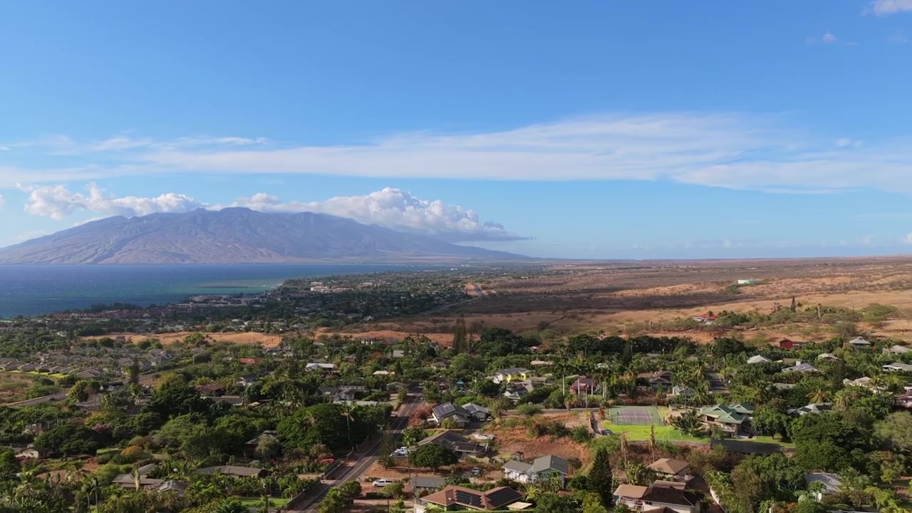 Video drone 4K a 60fps che si innalza da una lussureggiante vegetazione tropicale nel quartiere di Maui Meadows vicino a Wailea. Il volo verso l'alto rivela ampie proprietà immobiliari dell'isola, viste costiere e montagne lontane.