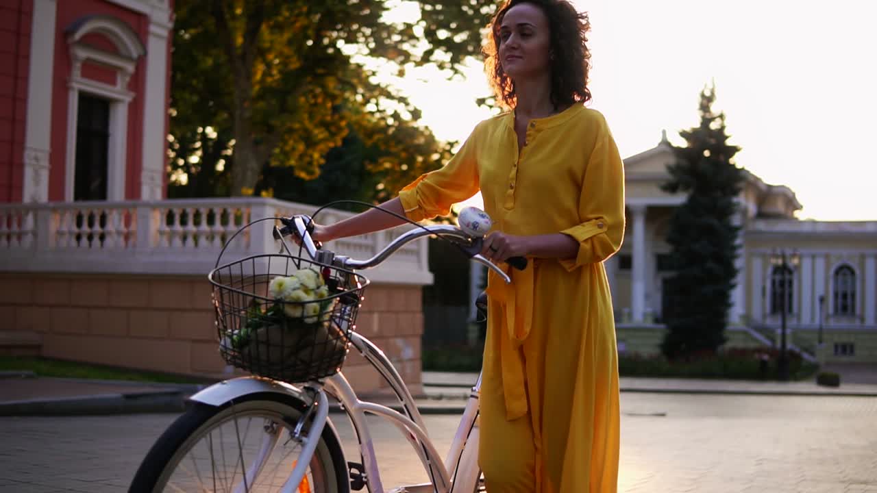 hermosa mujer joven en un largo vestido amarillo caminando durante el amanecer sosteniendo su manillar de bicicleta de la ciudad con flores en su