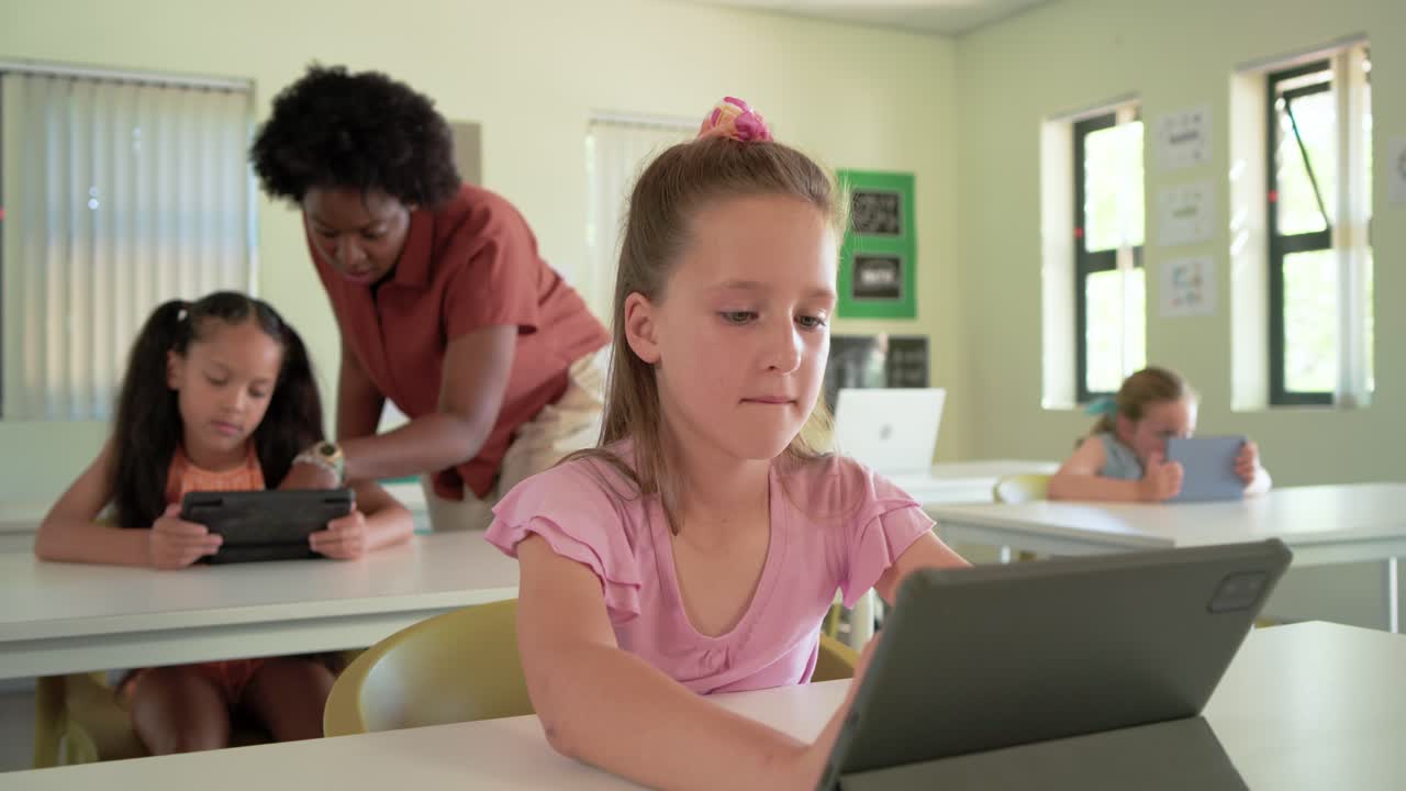 Classe eterogenea con l'insegnante che si avvicina alla scrivania, aiutando la ragazza al centro davanti che tocca il tablet e sorride.