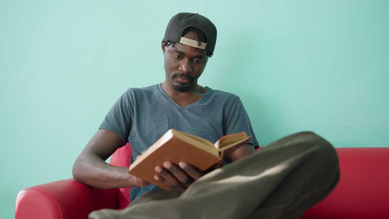 Nahaufnahme eines Mannes in einem lässigen Hemd und einer Mütze, der mit gekreuztem Bein auf einem roten Sofa sitzt, aufmerksam ein Buch liest und in einer hellen Innenumgebung mit türkisfarbener Wand auf eine neue Seite umblättert