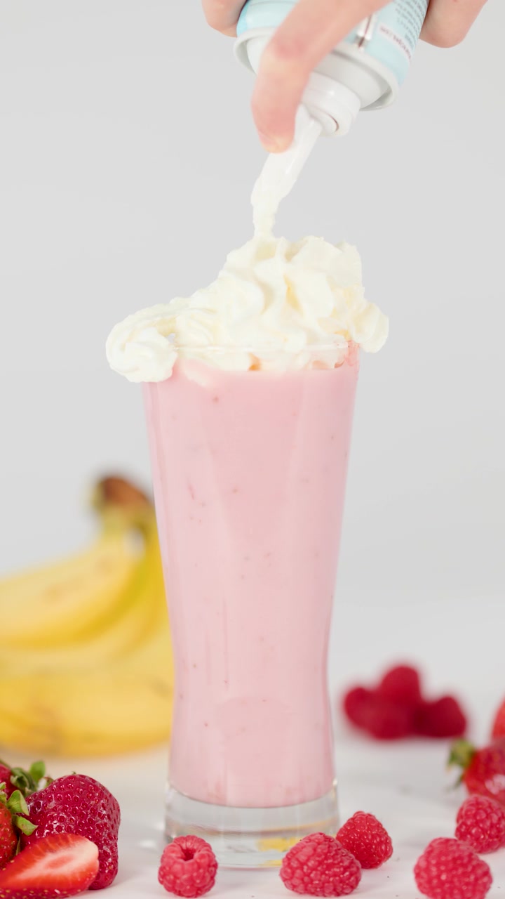 Hand tops pink smoothie with whipped cream, surrounded by fresh fruit, in bright studio lighting.