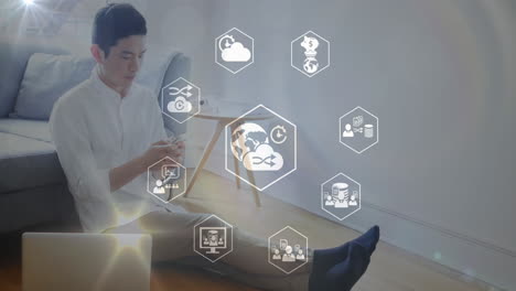 animación de iconos en hexágono sobre hombre asiático sentado en el suelo con computadora portátil y usando teléfono celular