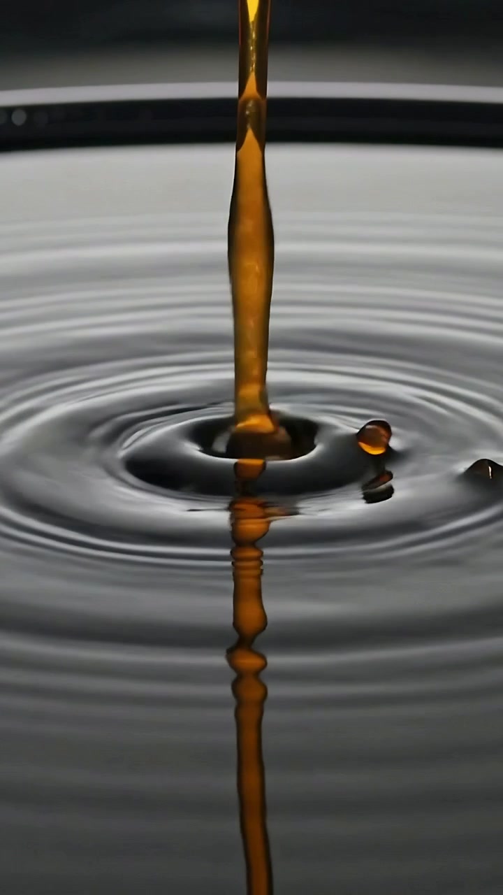 Vídeo vertical: Fluxo âmbar caindo e atingindo uma poça escura sobre a mesa, criando ondulações e uma pequena gota.