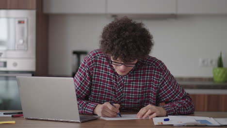 Estudiante-Masculino-De-Pelo-Rizado,-Un-Joven-Atractivo-Con-Gafas-Está-Estudiando-En-Casa-Usando-Una-Computadora-Portátil-Escribiendo-En-Un-Cuaderno.-Estudiante-Universitario-Usando-Una-Computadora-Portátil-Viendo-Un-Seminario-De-Aprendizaje-En-Línea-A-Distancia