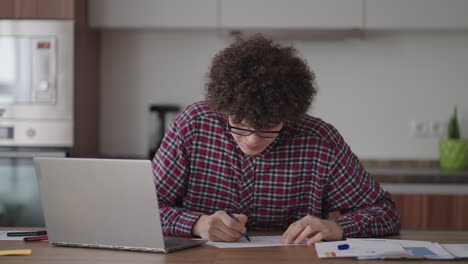 estudiante de cabello rizado atractivo joven con gafas está estudiando en casa usando una computadora portátil escribiendo en un cuaderno. estudiante universitario usando una computadora portátil viendo un seminario de aprendizaje en línea a distancia