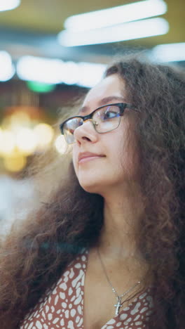 dama con cabello rizado vintage y gafas sentada en el restaurante, mirando a su alrededor pensativo, suaves luces bokeh en el fondo borroso crean un ambiente cálido y acogedor
