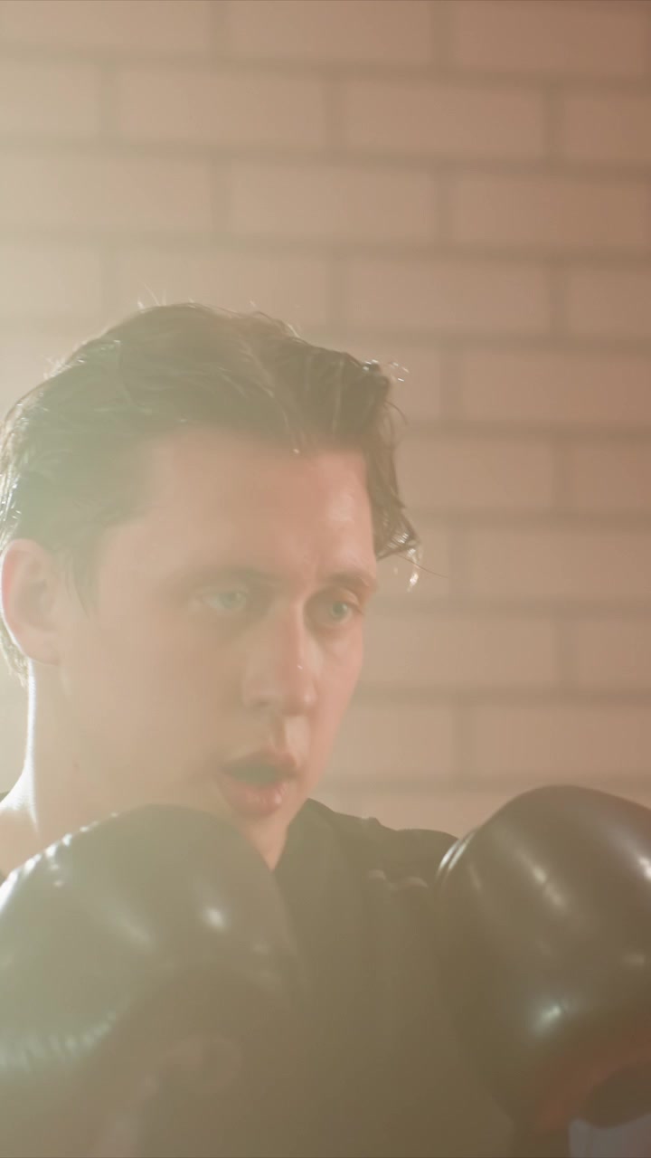 Close up of young man wearing black boxing gloves. Sweat visible on skin as man moves his head and upper body. Background features light colored walls with occasional brick