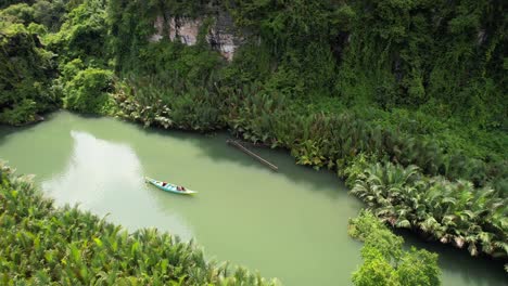 aerial view of a local boat floating down a green river in ramang ramang sulawesi surrounded by jungle, magroves, and palm trees