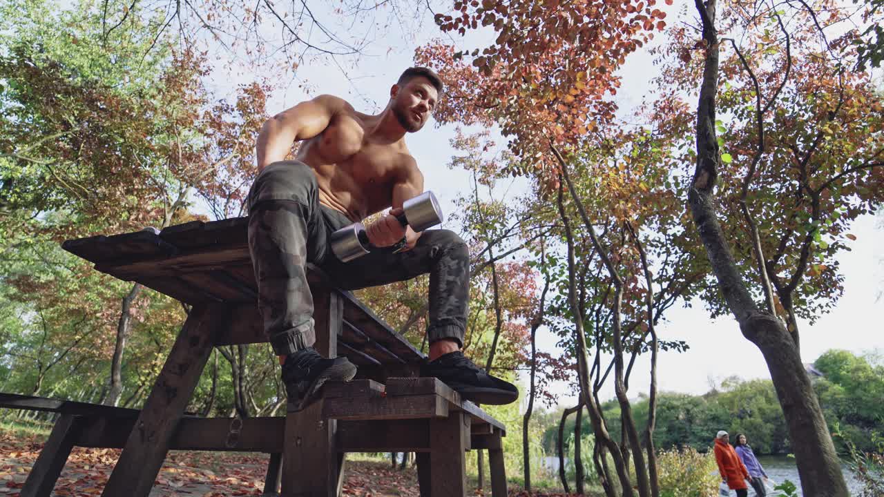 Shirtless sportsman lifting dumbbell outdoors. Strong athlete sitting on the table and pumping his arm muscles on autumn trees background. View from below.