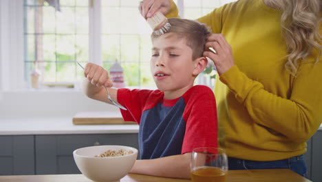 unhappy son at home eating breakfast cereal at kitchen counter as mother brushes his hair