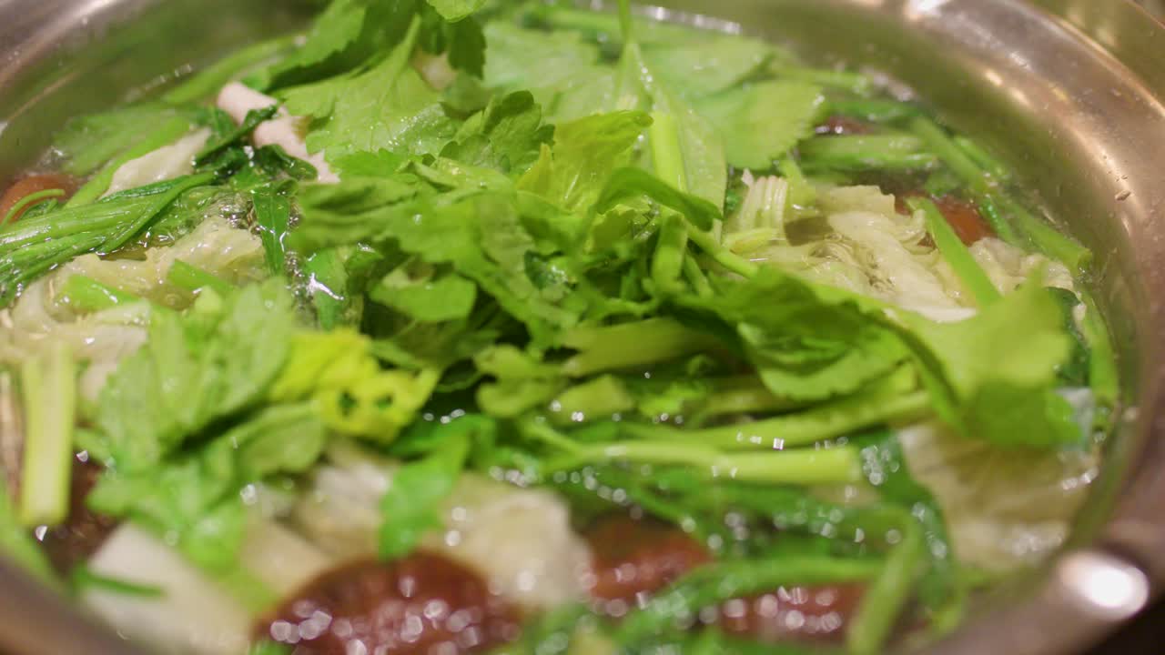 Assorted vegetables and sliced meat simmer in clear broth, overhead view, warm indoor lighting