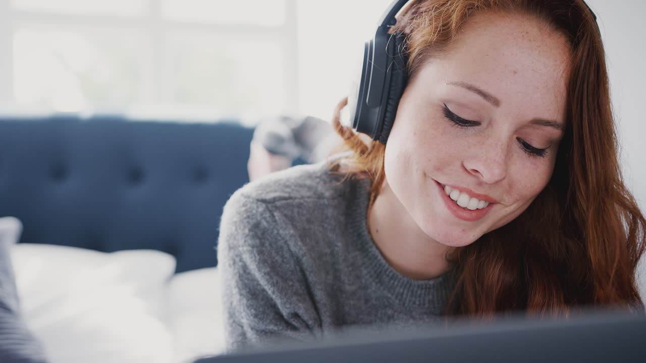 Kvindelig universitetsstuderende med hovedtelefoner arbejder på sengen i et delt hus med bærbar computer og mobiltelefon