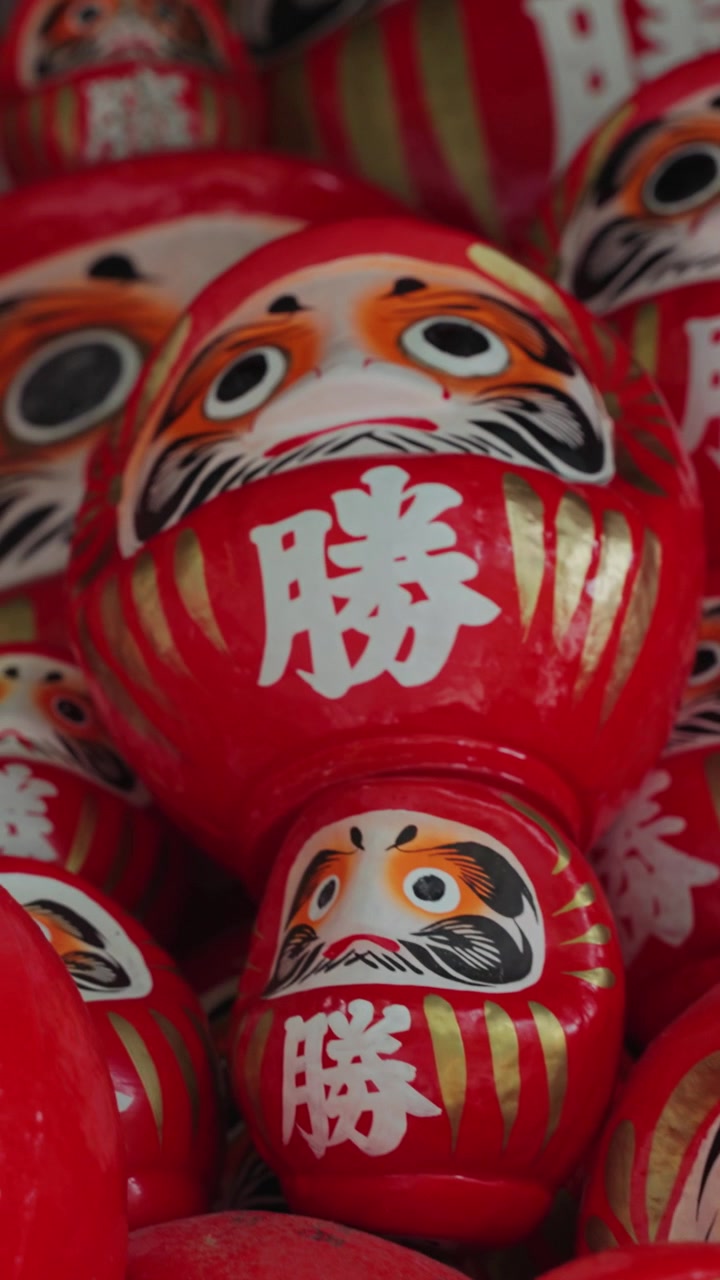 Video verticale che mostra file di bambole Daruma tradizionali al Tempio Katsuoji a Osaka, Giappone, che simboleggiano fortuna, determinazione, desideri e successo all'interno della cultura buddista giapponese.