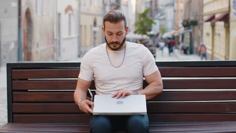 young man freelancer starts working on laptop sends messages makes online purchases on city bench