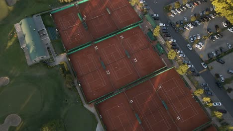 vista aérea de ángulo alto que revela canchas de tenis entre un campo de golf y un estacionamiento en un club deportivo al atardecer