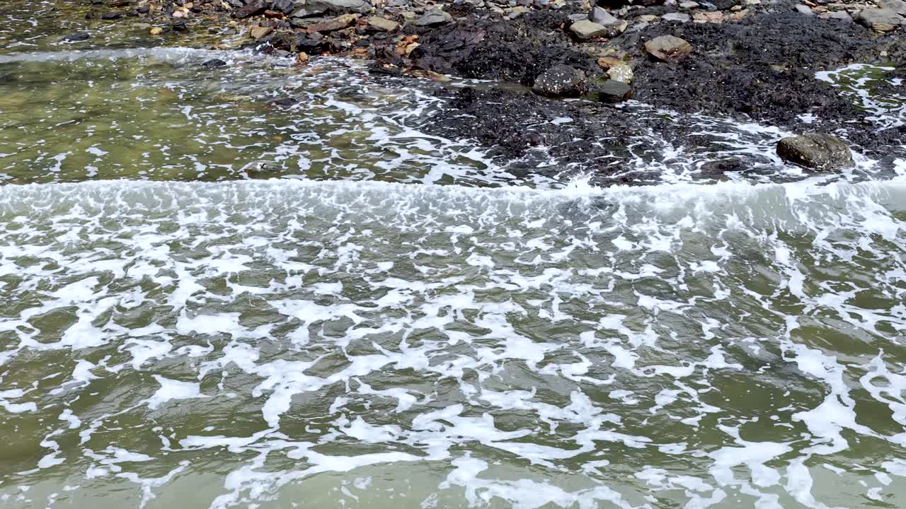 Foamy ocean waves break and recede over rocky coastal shoreline, daylight, steady wide shot